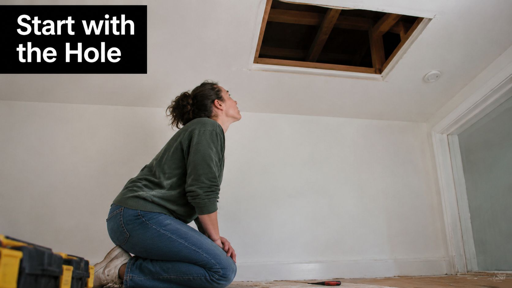 A woman kneeling on the floor while looking up at an open rectangular attic hatchway.